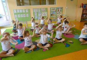 Dzieci siedzą skrzyżnie z uniesionymi nad głową rękami w których trzymają plastikowe butelki, wykonują skłony na boki.